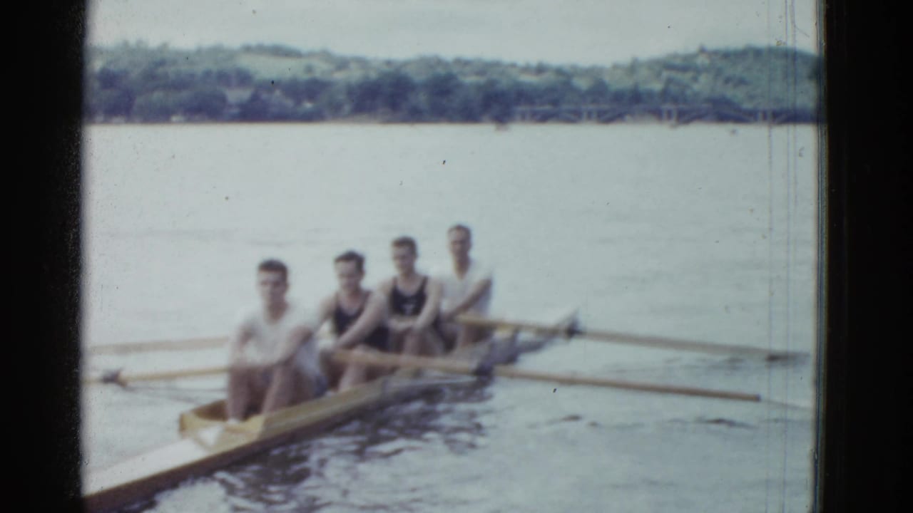 Four young men in a rowing shell appear on calm Boston-area water in 1951 home-movie footage, with a tree-lined shore behind them.