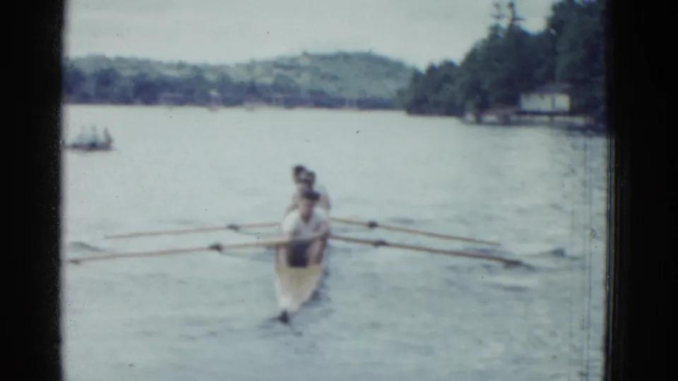 Seen from behind, the rowing shell travels away on calm water, with wooded hills and shoreline structures faintly visible in the distance.