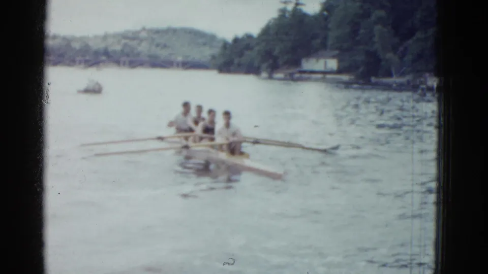 A 1951 Boston-area home-movie frame shows the crew rowing away across pale water, with small boats and trees in the background.