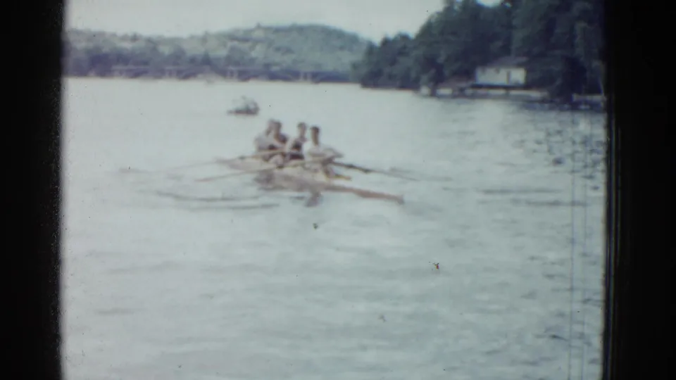 Soft 8mm frame from 1951 showing a four-man rowing shell on a Boston-area river with wooded banks behind it.