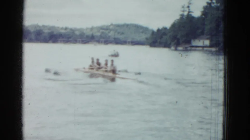 1951 Boston-area 8mm frame showing the rowing crew farther from camera on a calm river, with shoreline trees and hills visible.