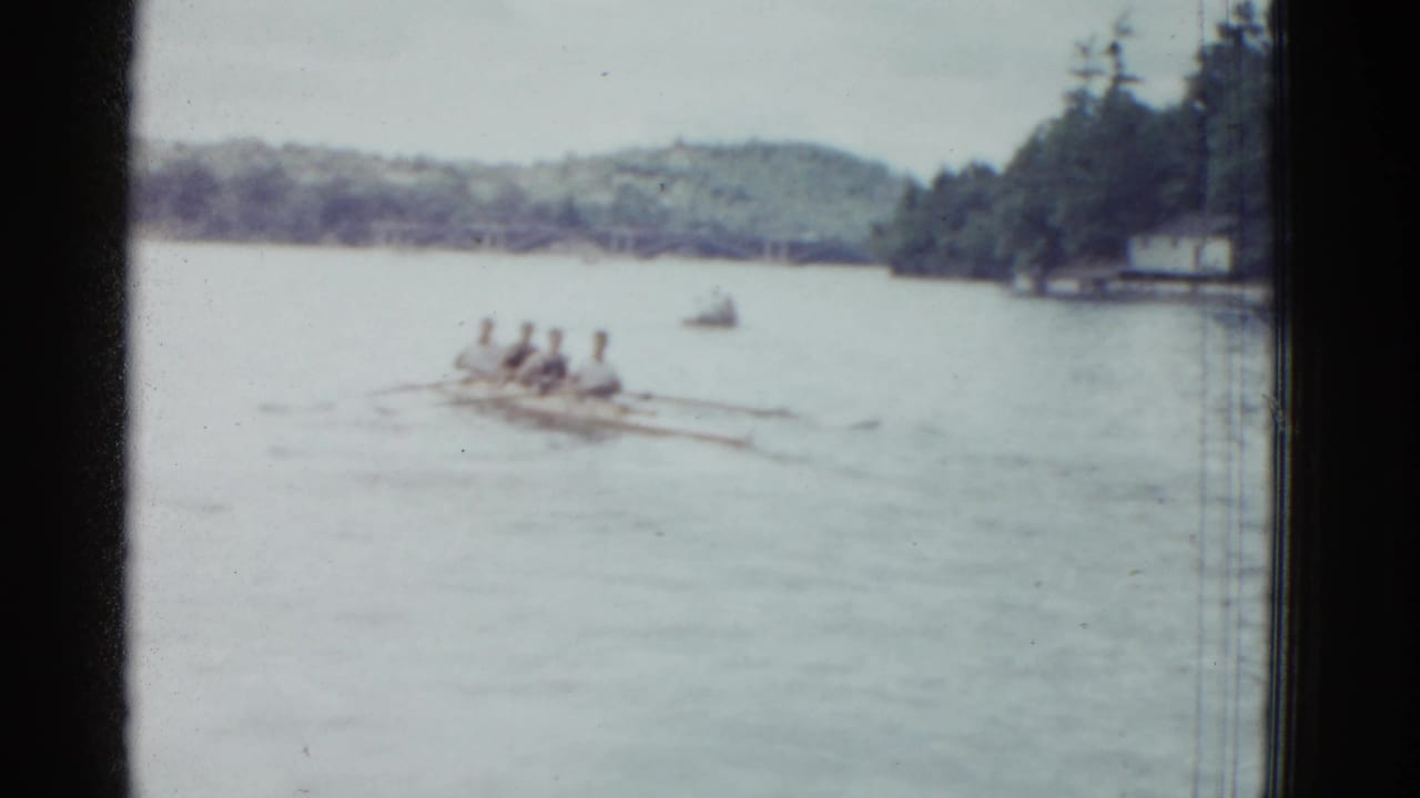 Four rowers in a long shell move across a broad river near a wooded shore.