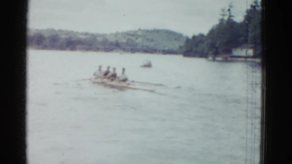 1951 Boston-area home-movie frame showing a distant rowing shell with four rowers on a wide river under a pale sky.