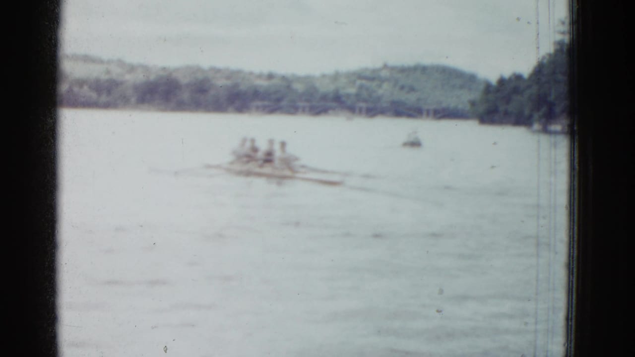 A rowing shell continues across open water with shoreline and small boats behind it.