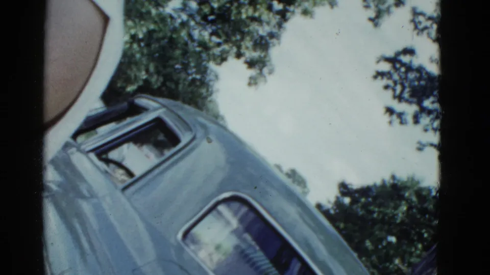 1951 Boston home-movie frame with a parked light blue classic car, leafy trees, pale sky, and visible 8mm film texture.