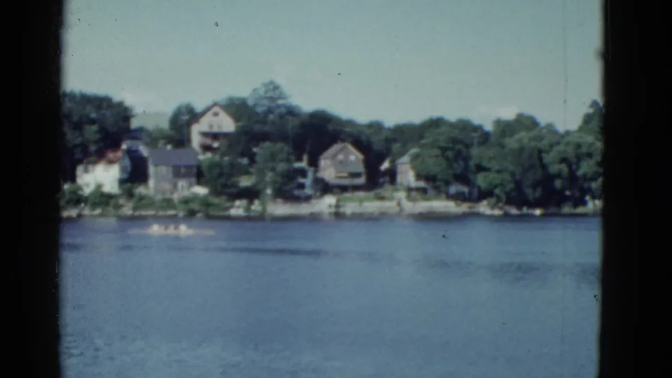 1951 Boston-area lakeside home-movie frame with broad water, tree cover, and houses across the shore.