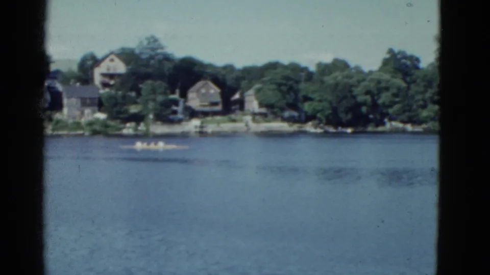 1951 Boston-area river scene with a small boat passing below homes set among trees on the far bank.