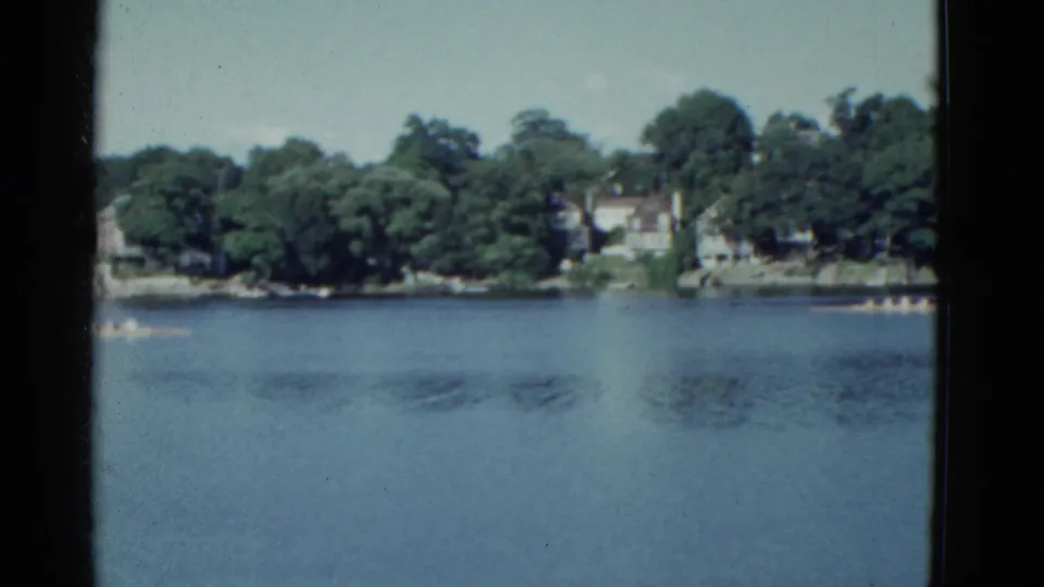 1951 Boston-area riverfront frame with the boat near the left edge and residential shoreline across the water.