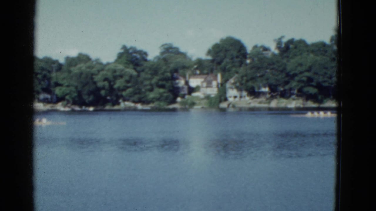 Distant canoe teams race across a blue lake with trees and houses on the far shore.
