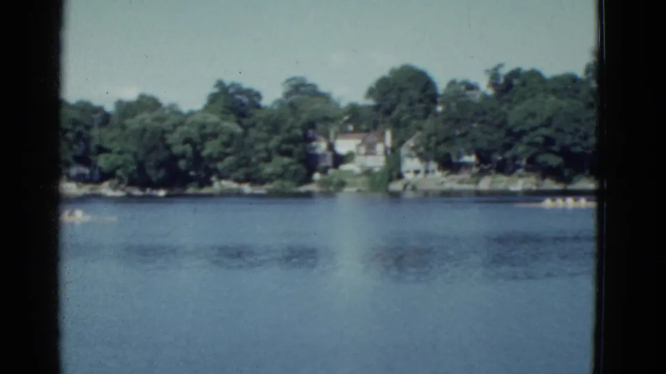 1951 Boston-area 8mm view of canoe teams moving across calm blue water, with wooded shoreline and houses in the background.