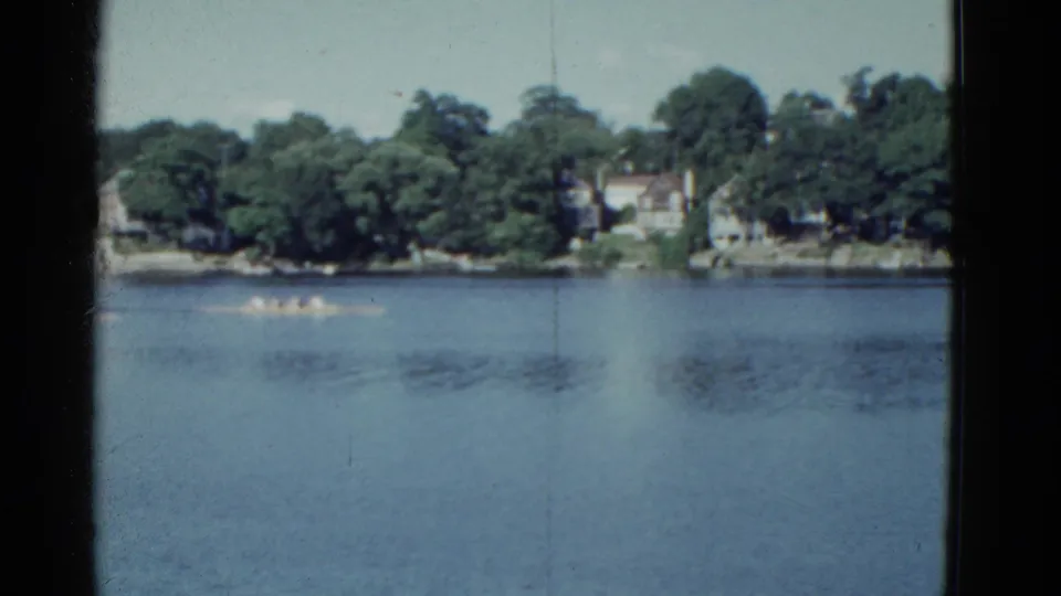 Distant 1951 Boston-area lake scene showing canoe racers in formation, framed by trees and shorefront houses.