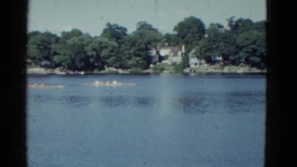 1951 Boston-area 8mm frame with canoe teams crossing blue water below a leafy residential shoreline.