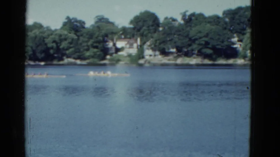 Boston-area 1951 home-movie frame of canoe racers on a lake, seen from across the water with wooded homes beyond.