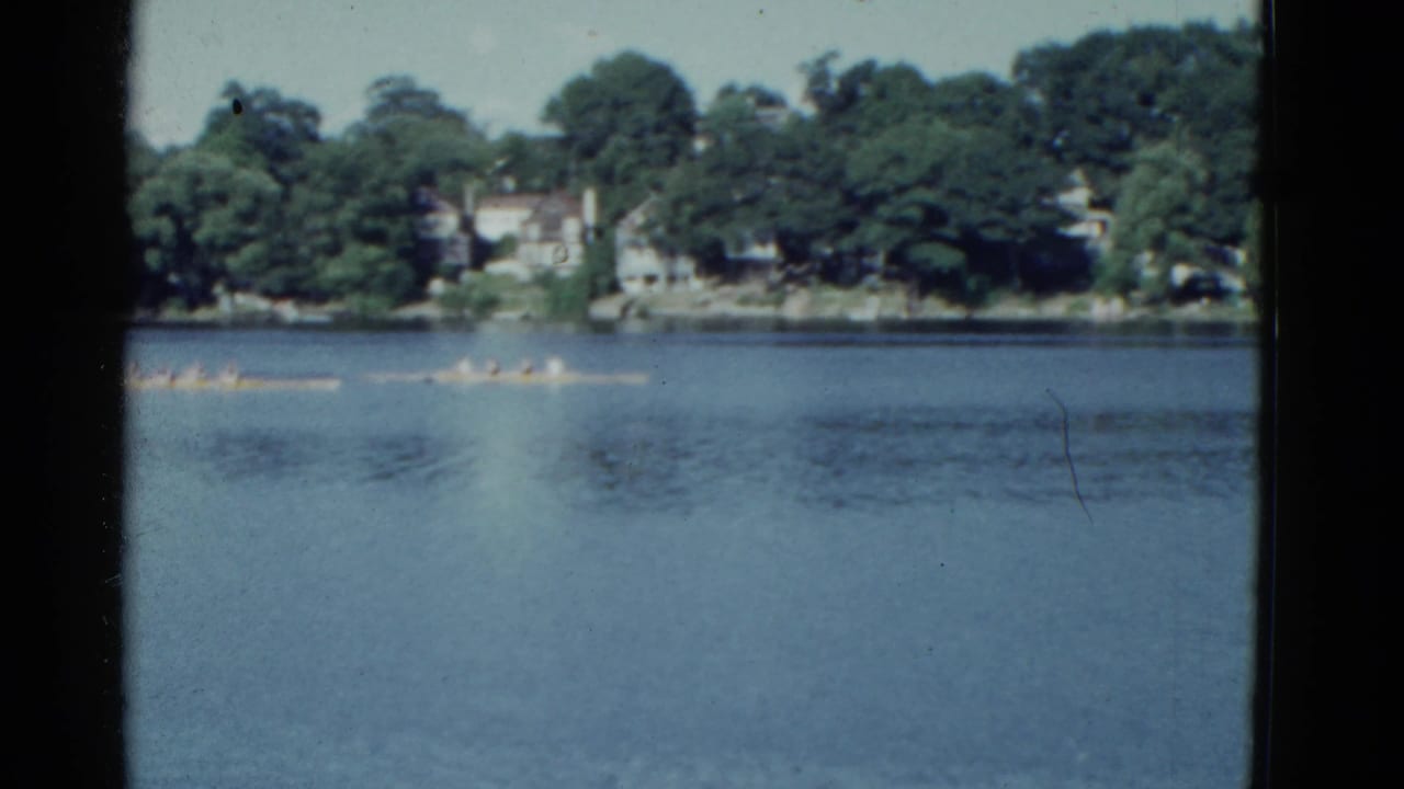Rowing shell crossing calm water with a tree-lined shore behind it.