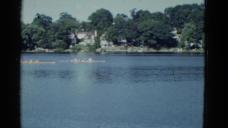 Distant rowers move across calm water in 1951 Boston-area home-movie footage, with trees and houses along the far shore.
