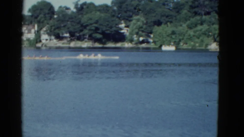 The rowing crew remains in the middle distance on blue water, with leafy shoreline and light-colored homes in the background.