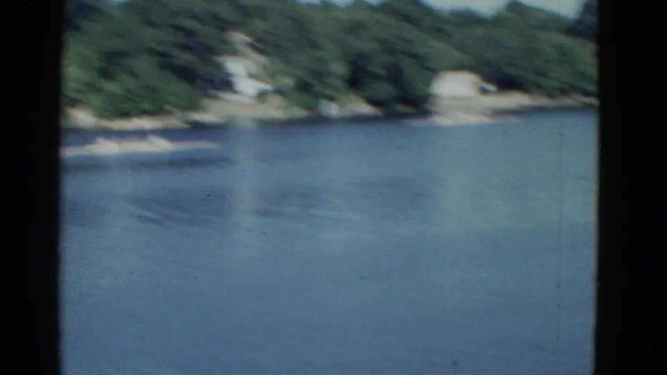 1951 Boston-area 8mm frame showing a calm lake, wooded shore, distant houses, and small rowing craft near the far bank.