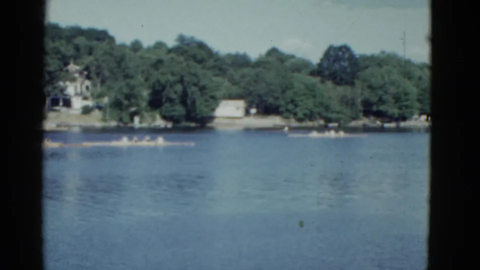1951 Boston-area home-movie frame showing multiple rowing shells spread across calm water near trees and shoreline structures.