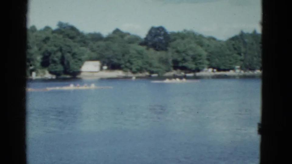 Boston-area 1951 8mm frame of rowing shells on a lake, backed by dense summer trees and a small light-colored shoreline building.