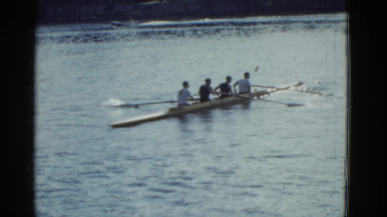 1951 Boston 8mm frame showing a four-person rowing shell on open river water, seen from a distant home-movie viewpoint.