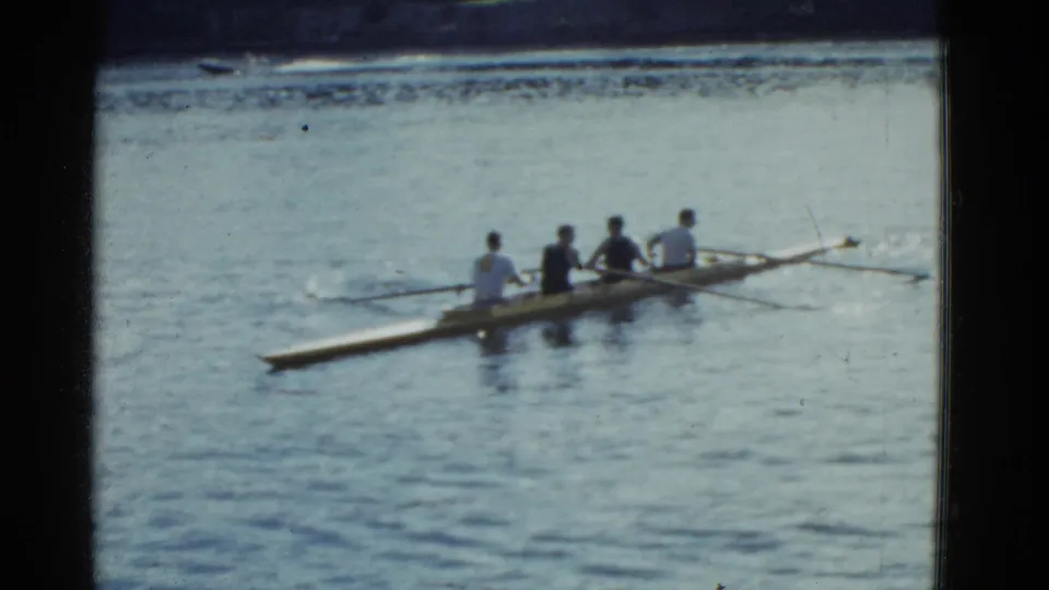 A 1951 Boston home-movie frame shows the shell from the side, with four rowers seated in sequence and oars reaching outward.