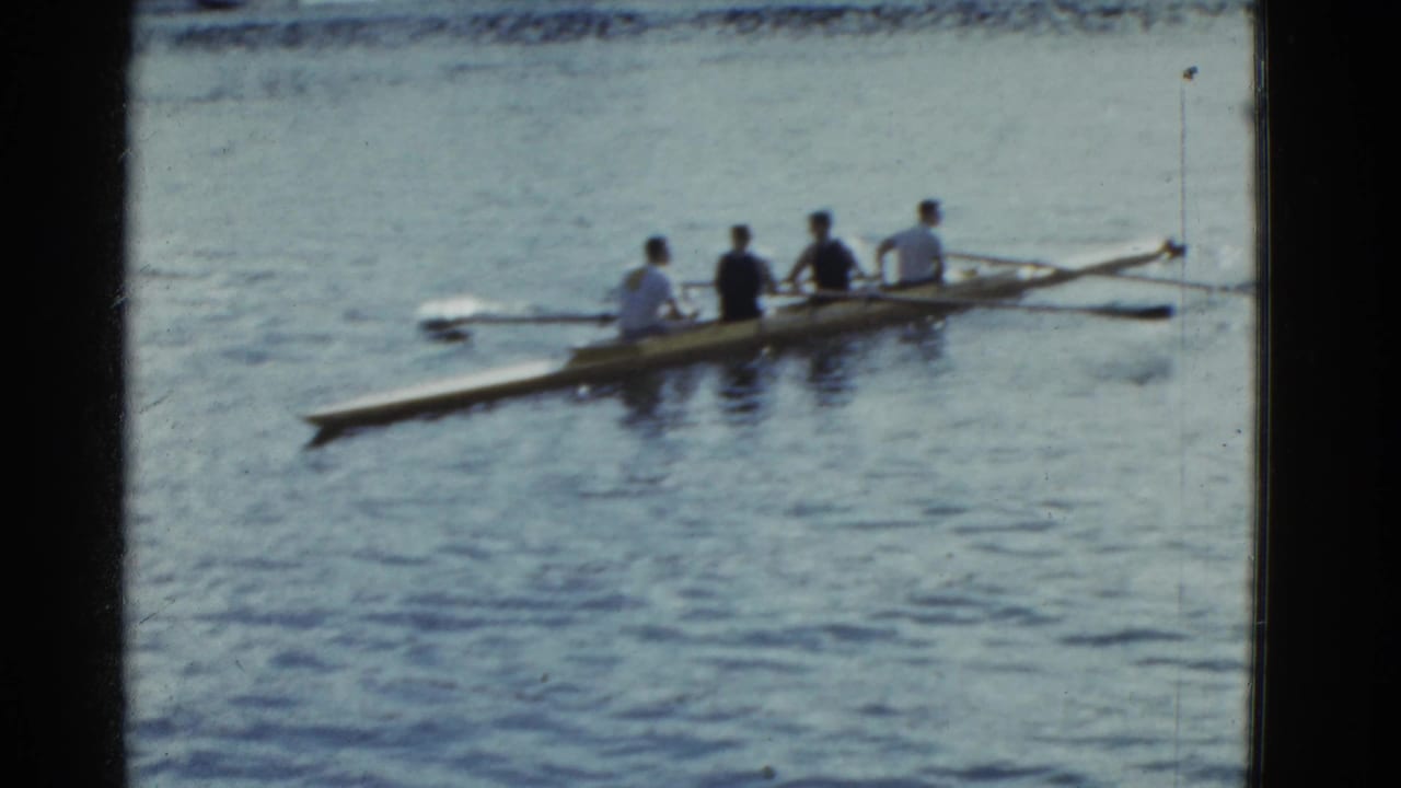 1951 Boston-area 8mm frame showing four men rowing a narrow shell across calm water in bright daylight.