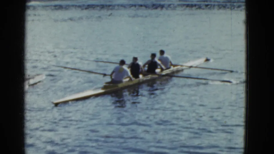 Four rowers in a narrow shell pass through the frame on calm water in 1951 Boston-area home-movie footage.