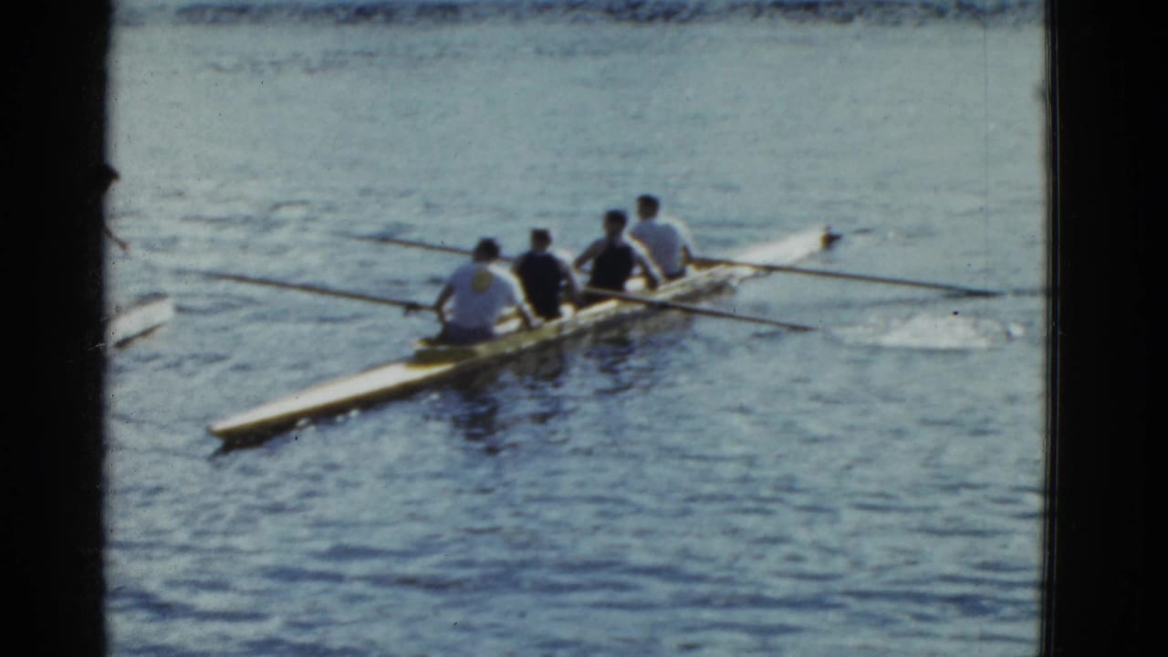 Four-person rowing shell on blue water with oars extended.