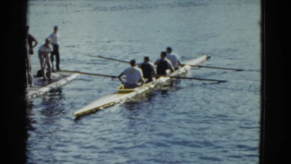 A 1951 Boston-area rowing crew continues past a nearby support boat and standing figures.