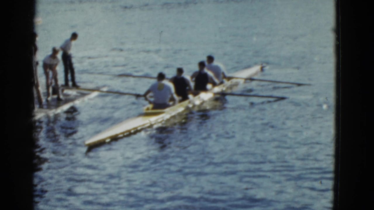 Rowing team in a long shell beside a dock on calm water.