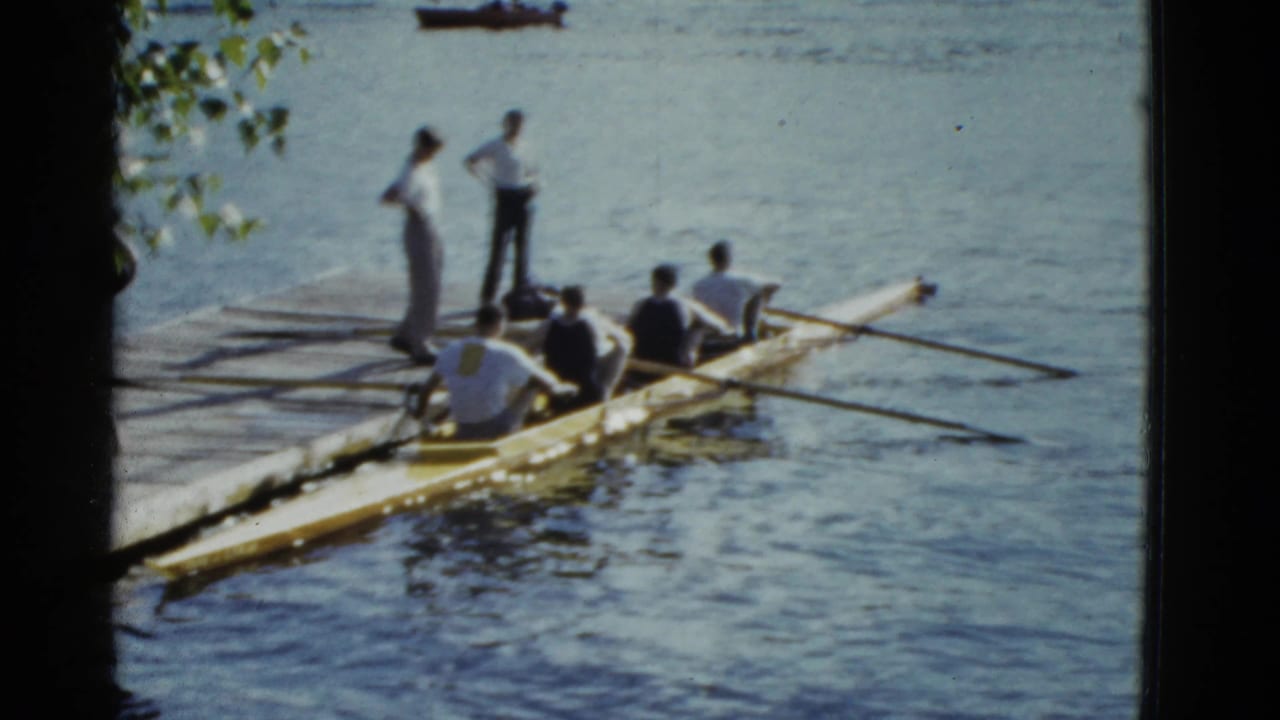 Rowing shell beside a dock with men preparing oars on a calm river.
