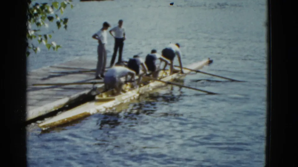 Adult rowers in a 1951 Boston river scene adjust positions in a long shell while two men stand on the dock.