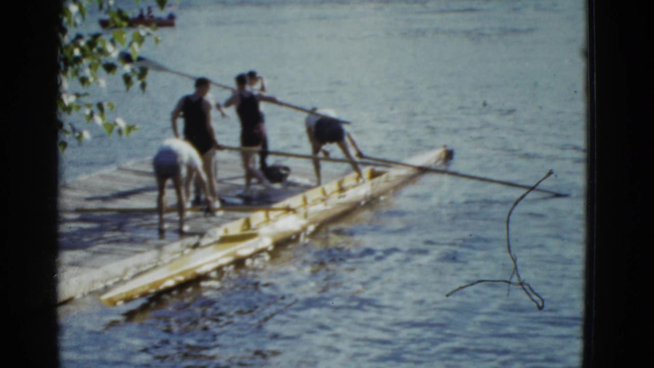 1951 Boston home-movie frame showing men handling a yellow rowing shell and oars from a wooden dock.