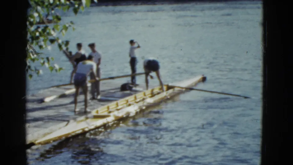 1951 Boston frame of men preparing or retrieving a rowing shell, with oars extending over the water.