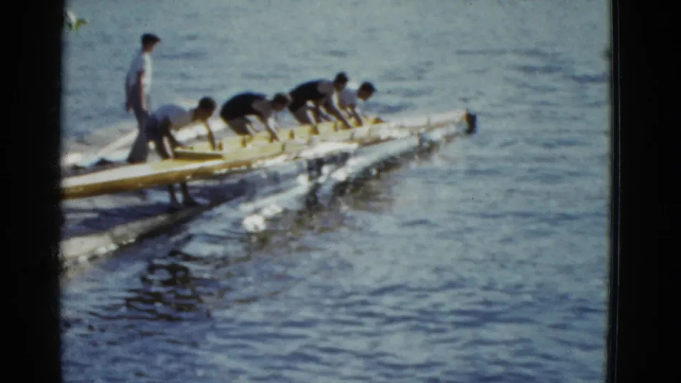 A 1951 Boston home-movie frame shows men leaning over a yellow rowing shell near rippled water and dock edge.