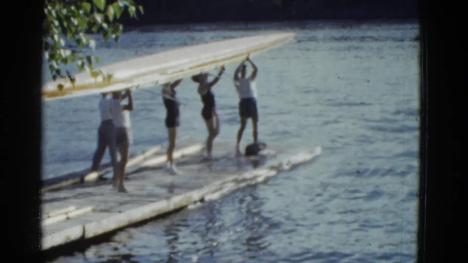 Several men raise a rowing shell overhead on a dock, a brief 1951 Boston home-movie moment of coordinated launch preparation.