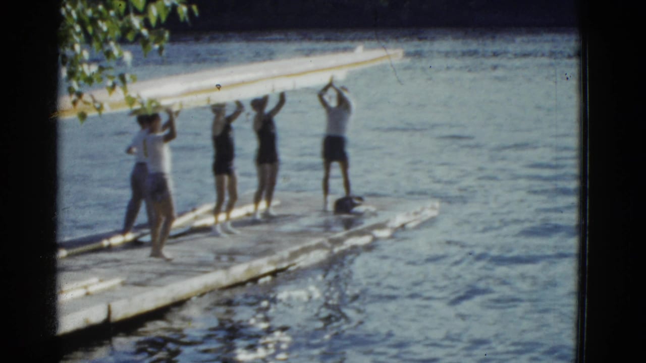 Young men stand on a dock lifting a long boat beside lake water.