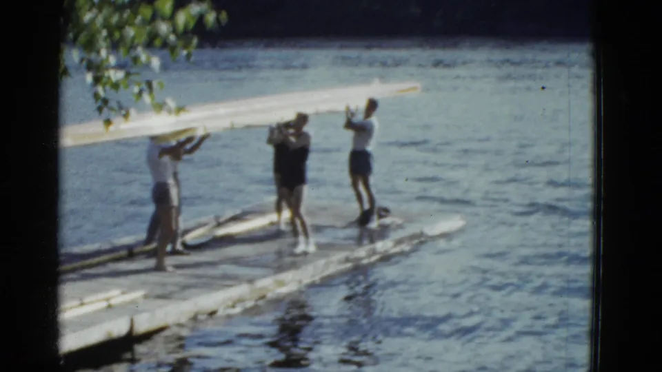 Young men carry a long wooden boat along a lakeside dock in 1951 Boston-area 8mm footage.