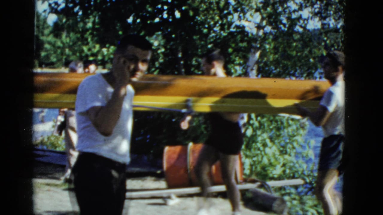 A man in a white T-shirt stands by a long wooden boat as others carry it near the water.