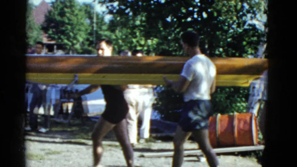 Men in swimwear carry a long wooden boat past red drums and trees along a lakeside path in 1951 Boston home-movie footage.