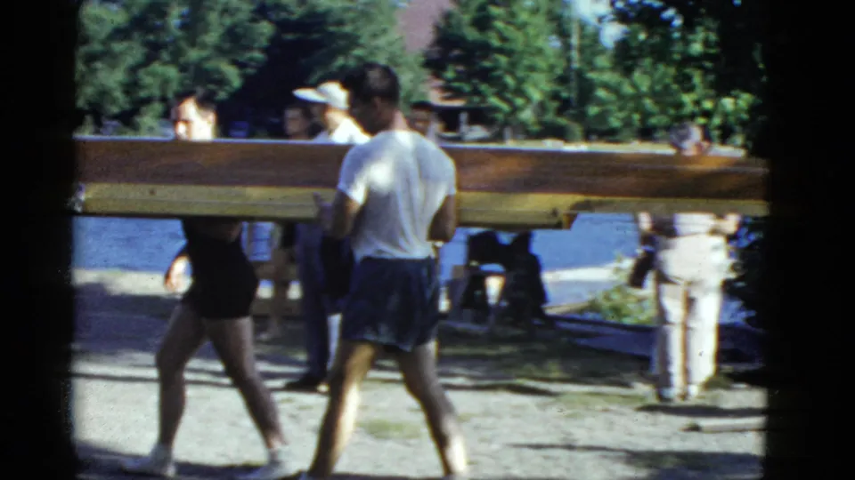 Men carry a wooden boat through a shaded lakeside area, with water and trees visible in this 1951 Boston home-movie frame.