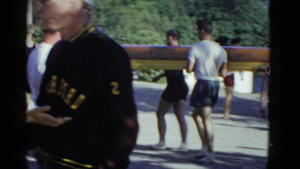 A man in a Harvard sweater stands near a carried rowing shell.