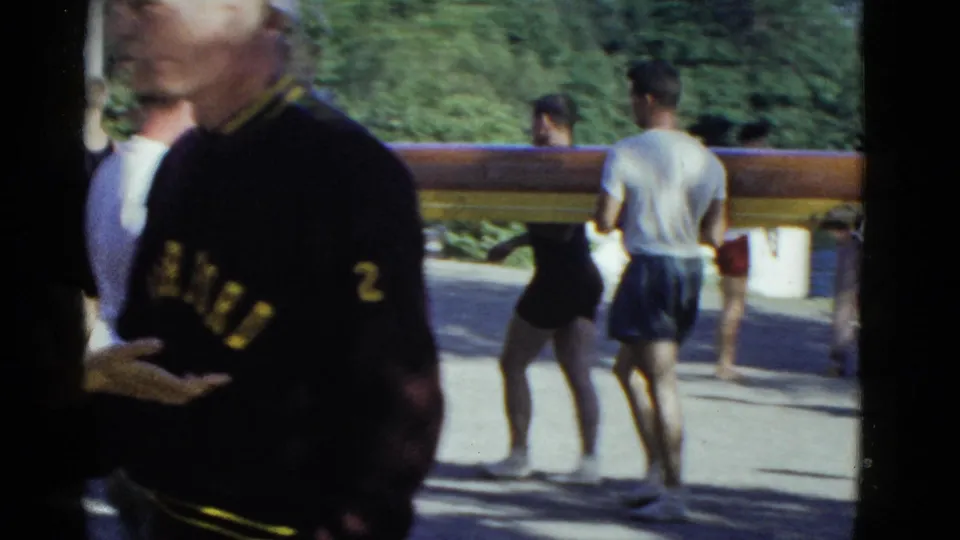 A man wearing a dark Harvard sweater with number 2 is seen beside a long rowing shell in the Boston 1951 footage.