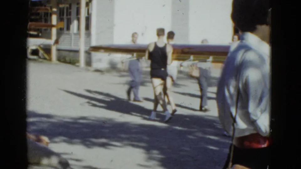 Men in athletic wear carry the long wooden shell through a bright Boston-area rowing path, with bystanders nearby.