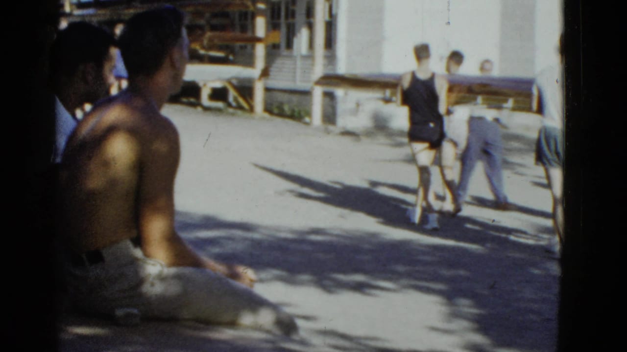 1951 Boston-area home-movie frame showing adults near a lakeside camp or boathouse, with rowing shells and swimmers in view.
