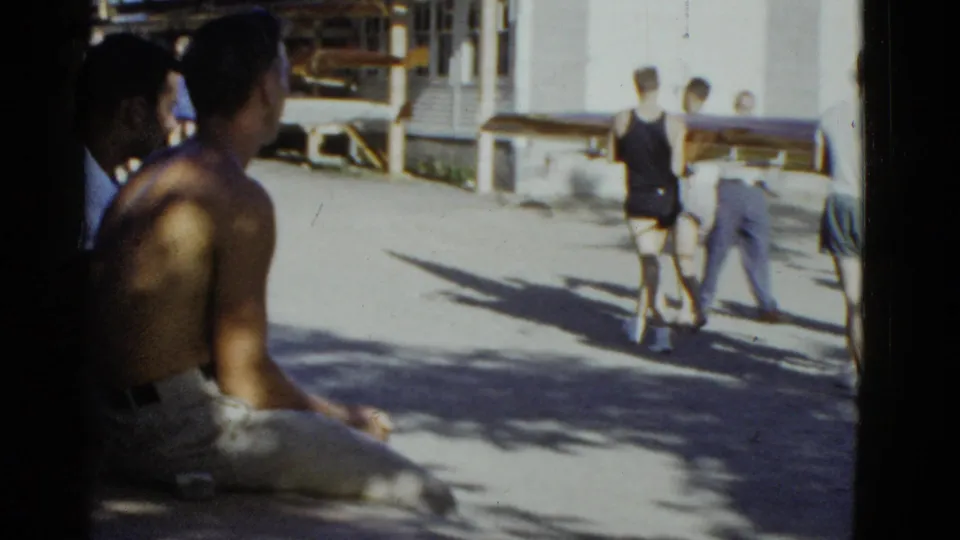 1951 Boston-area home-movie frame showing adults near a lakeside camp or boathouse, with rowing shells and swimmers in view.