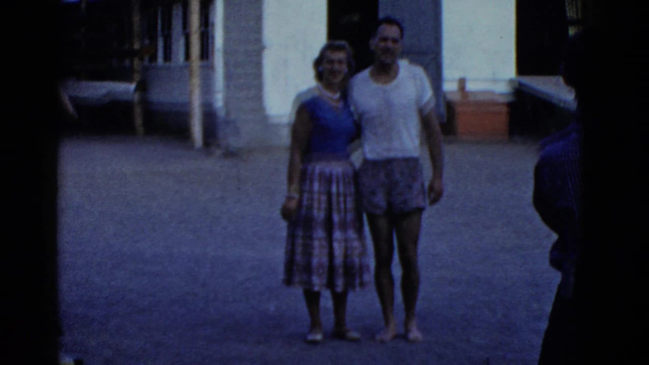 A woman and man stand together outdoors near a camp building and rowing equipment.