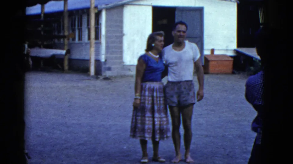 1951 Boston-area home-movie frame of a couple posing arm in arm, with camp structures and shaded equipment behind them.