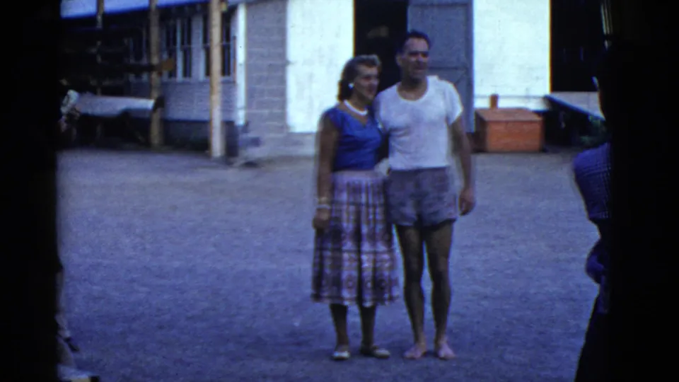 1951 Boston-area frame showing a couple standing side by side near a lakeside camp building, with rowing equipment partly visible.