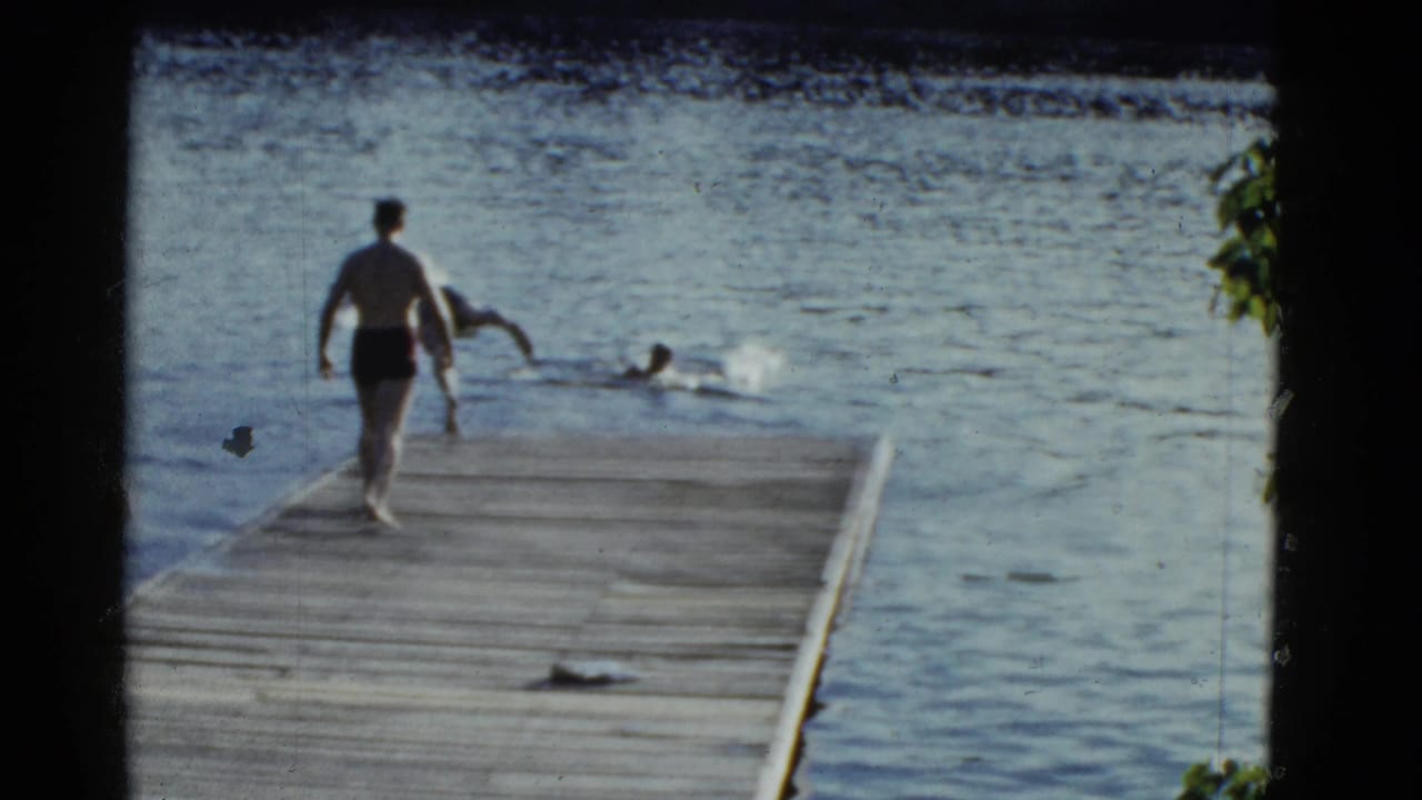Man in dark swim trunks walks along a wooden pier while swimmers move in the lake.